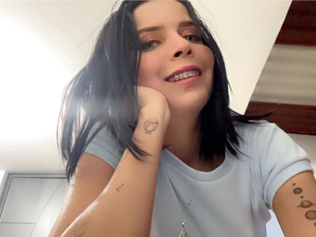 KaylinJulithe tem cabelo preto, uma camisa azul clara, tatuagens no braço e está sorrindo, com a mão no rosto, mostrando os dentes. Seu nariz é ligeiramente arrebitado..