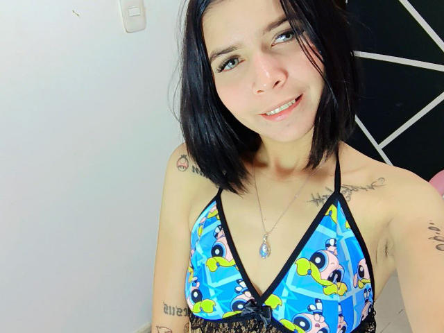 KaylinJulithe, con cabello negro corto, sonriendo, vestida con un top de bikini azul con estampado de dibujos animados, mostrando pequeños senos, tatuajes en los brazos y un collar de plata. Está tomando una selfie..