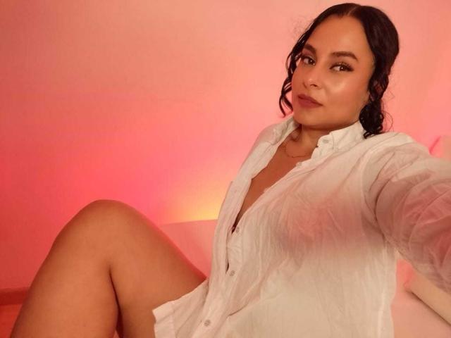 LunaSoler, com seios médios, vestindo uma camisa branca de botões, sentada com as pernas levantadas, fundo iluminado em rosa, sorriso suave, cabelo escuro ondulado, pele clara, selfie casual..