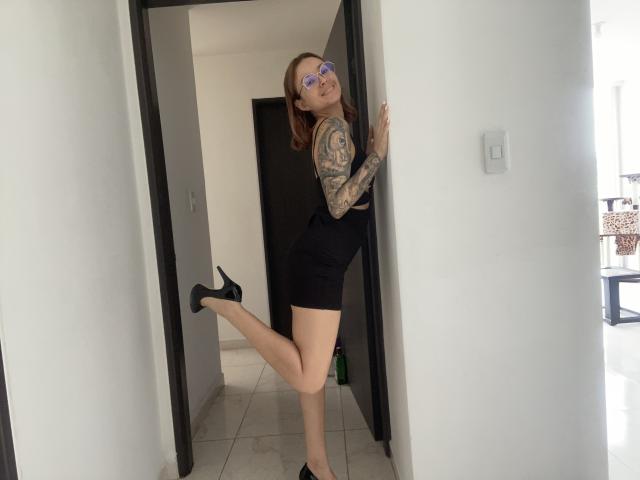 SarahRousen with tattoos on her arm, wearing glasses, black high-waisted shorts, and black heels, standing in a doorway, leg raised, showing her bare ass and smiling.
