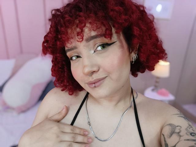 TracieLowe with curly red hair, green eyeliner, nose ring, lip piercing, silver chain, tattoo on right arm, black halter top, and a hand on her chest, smiling softly.