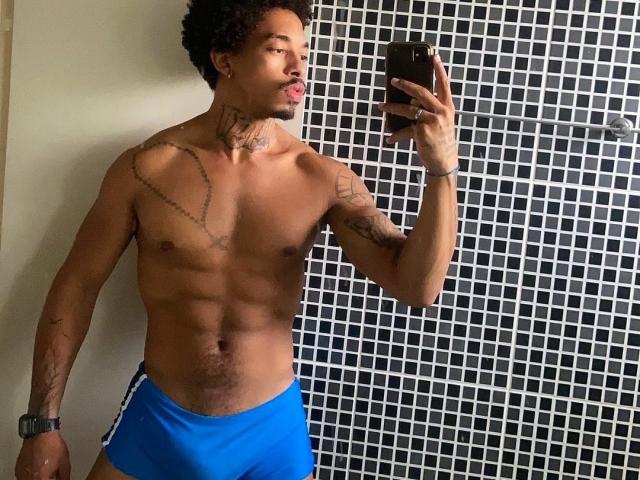 BethenCurtDotado with curly hair, tattoos, and a blue bulge, taking a mirror selfie, tongue out, wearing blue shorts, black watch, holding a phone.