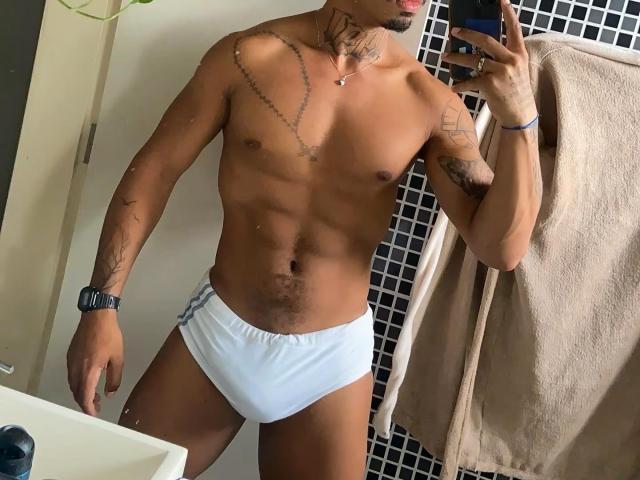 BethenCurtDotado, muscular with tattoos, wearing white boyshorts, has a flat stomach and is taking a mirror selfie with a phone. He has a watch on his left wrist.