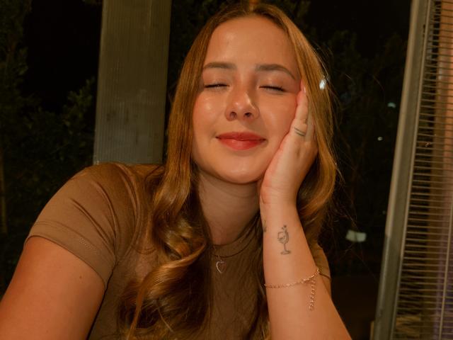 NatSorian met gesloten ogen, lachend, met haar hoofd rustend op haar hand, met een kleine tatoeage van een lieveheersbeestje op haar pols, gekleed in een bruin shirt en een ketting..