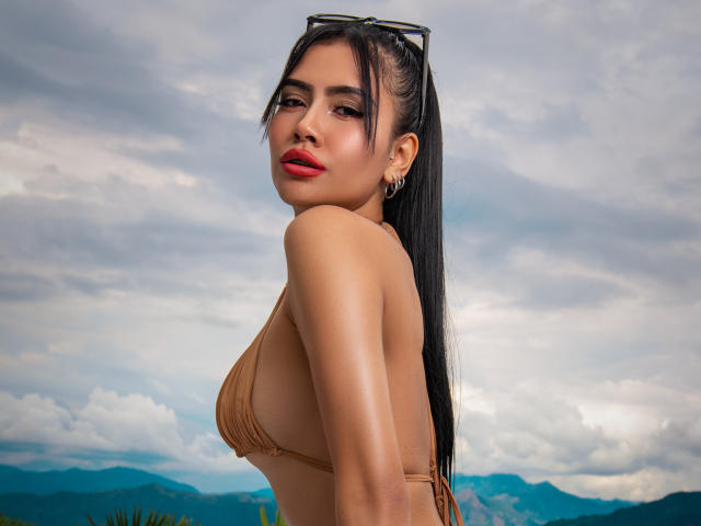 TinnyEngel com top de biquíni bege, mostrando pequenos seios, cabelo preto comprido em rabo de cavalo, batom vermelho, brincos, óculos de sol na cabeça, olhando para trás, sob um céu nublado, com montanhas ao fundo..