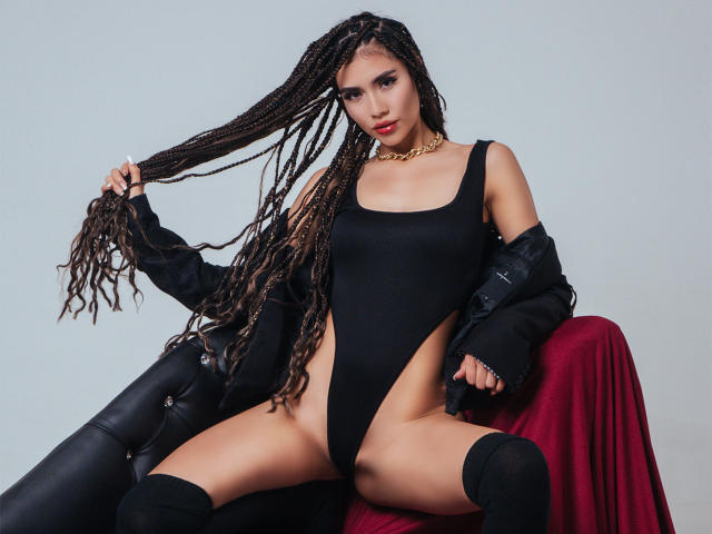 Een TinnyEngel met lange vlechten, gekleed in een zwart geheel, met een jas uitgetrokken, een gouden ketting en zwarte sokken, zittend op een rode stoel, waarbij haar vagina zichtbaar is door de hoge uitsparingen aan de zijkanten..