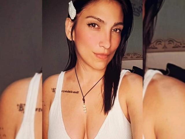 MicaelaJones with black hair, white flower in hair, white tank top, necklace, tattoos on shoulder, reflection in mirror, brown eyes, slight smile, cleavage visible, medium skin tone.