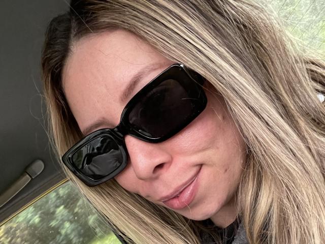 JennyLawry, met blond haar, draagt zwarte zonnebril en glimlacht lichtjes, waarbij een beetje van haar neus en lippen zichtbaar zijn. Ze zit in een auto met een groene buitenkant. Haar gezicht is dichtbij de camera..