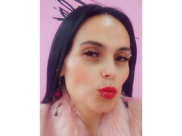 CharlotteCrofort with black hair, red lipstick, and a nose piercing, pouting with a pink feathered jacket, wearing pink earrings, has a shiny forehead, and a cute, sexy look.