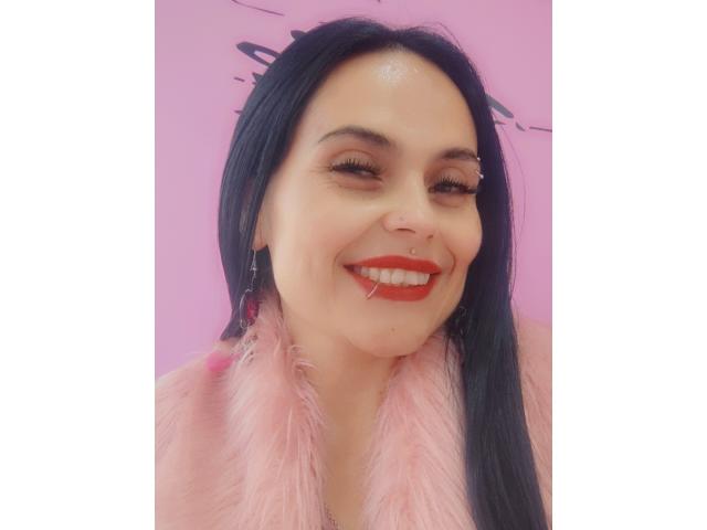 CharlotteCrofort with black hair, red lipstick, and pink feathered top, smiling with eyes closed, showing off her shiny nose and long eyelashes. She's got a cute, sexy face.
