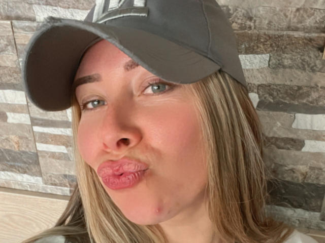 JennyLawry with puffy pink lips, blonde hair, wearing a grey cap, blue eyes, light skin, slight zit on cheek, making a puckering face.