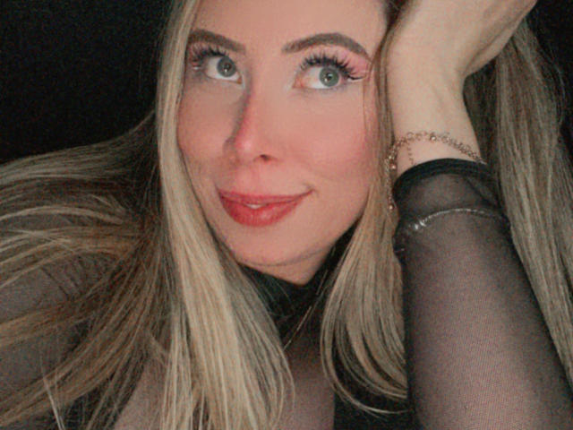 JennyLawry with blonde hair, green eyes, and pink lipstick, smiling at the camera, wearing a black sheer sleeve, hand resting on her head. Her tits are not visible.