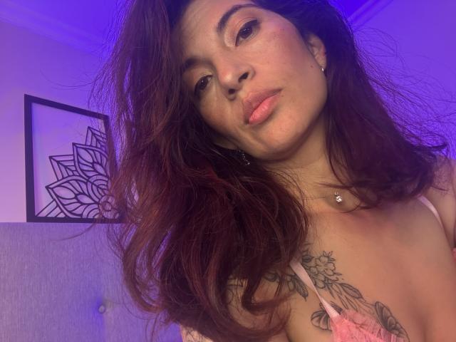 LaurentColin with messy brown hair, pink lips, and a tattooed chest, wearing a pink lace top, looking at the camera with a slight pout, purple lighting in background.