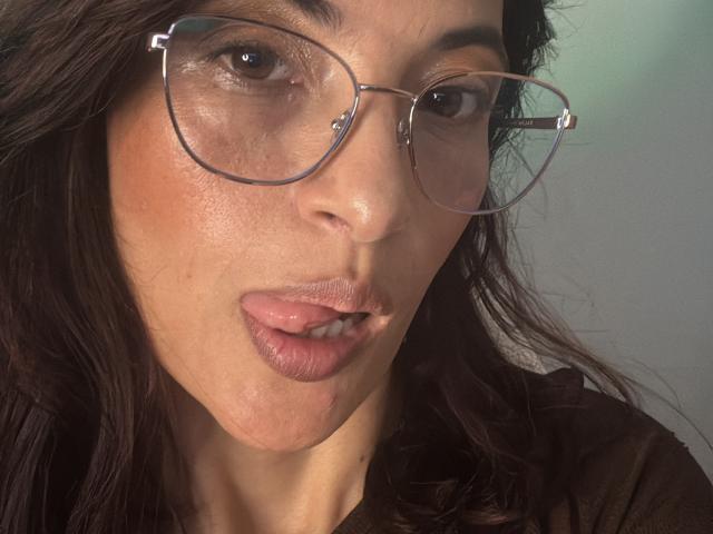 LaurentColin tem a pele morena, cabelo castanho ondulado e está com a língua rosa para fora, usando óculos. Seus lábios estão ligeiramente entreabertos..