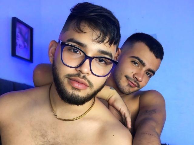 Two shirtless RandomGairysGuys with dark hair and beards, one with glasses and a gold necklace, the other with a tattooed arm, standing closely, blue-lit background.