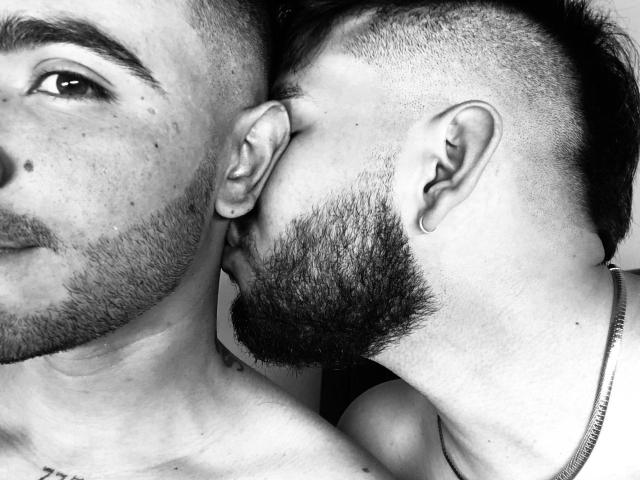 Two RandomGairysGuys, both with beards and shaved sides, kissing near the ear. One has a necklace, the other's face is partially visible. Black and white photo.