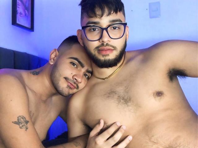 Two RandomGairysGuys, shirtless, in a bedroom. One has glasses, beard, chest hair, and a gold necklace, while the other has tattoos, a shaved head, and a tattoo on his arm.