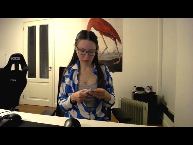JuliaStoneOne with glasses, blue and white patterned robe, gray top, looking down at phone, standing in room, next to black chair, white door, red bird painting in background.