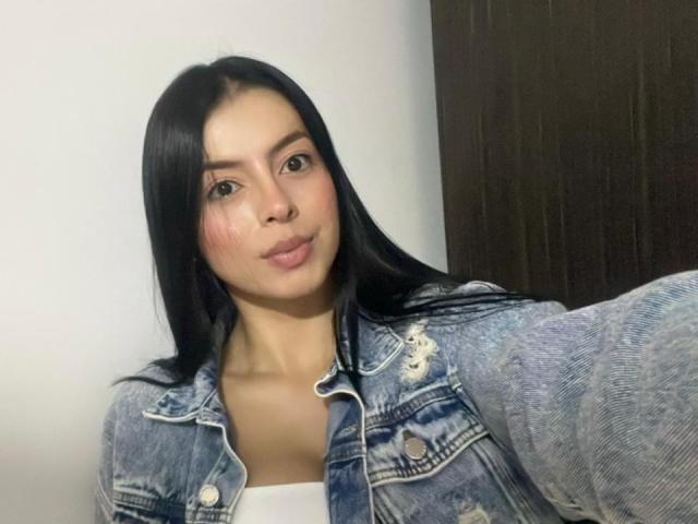 IsabellaJames with long black hair, wearing a blue denim jacket, puffy pink cheeks, and a white top, taking a selfie, showing her cute face and small, round nose.