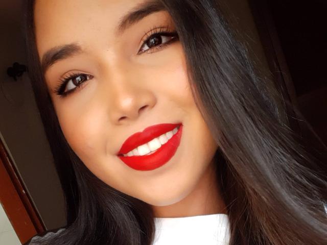 Heyly, una donna asiatica sexy con lunghi capelli neri, sorridente con un rossetto rosso brillante, sta sorridendo direttamente alla telecamera.