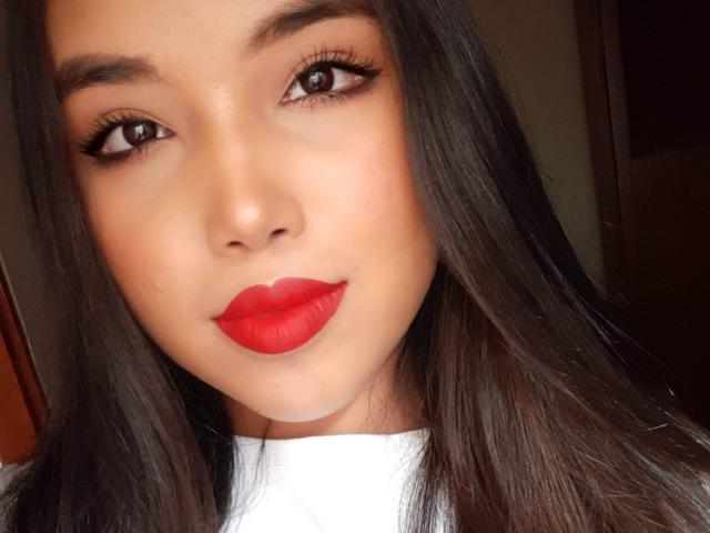 Heyly has bright red lipstick, black eyeliner, and long black hair, looking directly at the camera with a serious expression.