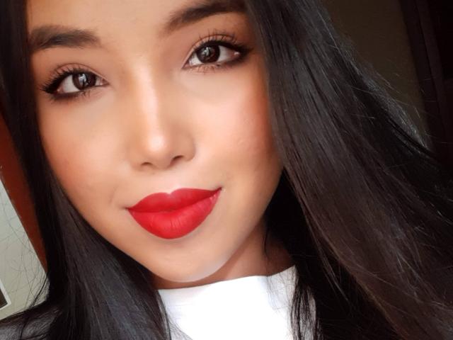 Le visage de Heyly est en gros plan, avec du rouge à lèvres rouge vif et des cheveux foncés et droits, montrant un sourire séduisant et un regard direct.