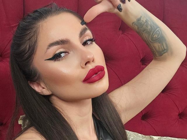 GoddessKaterina has dark hair, bold black eyeliner, red lipstick, and a tattoo on her arm. She's touching her forehead with one hand, showing black nails.