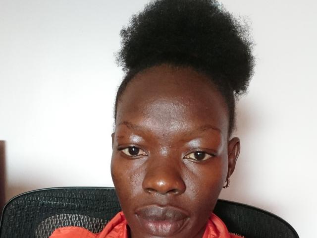 KrisJohanas has dark brown skin, small nose, and a tight Afro. She's wearing a red shirt, sitting in a black mesh chair, with a serious expression.