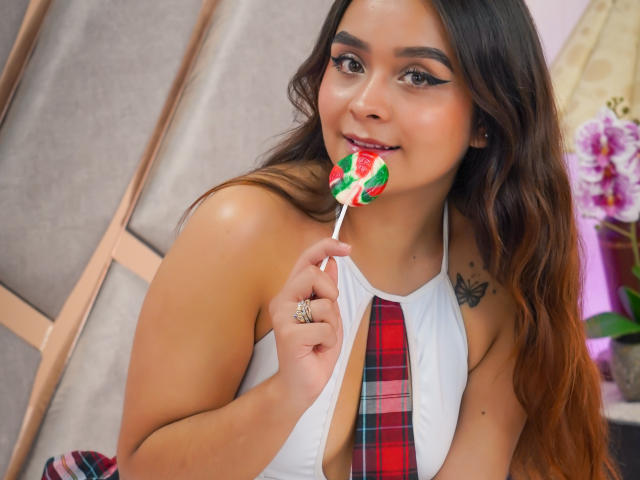 ViolaKaupp mit langen, braunen Haaren, einem weißen Top mit Halter, einer roten Karokrawatte, die an einem Zuckerrohr lutscht, ihren Ring in der Hand, einem Tattoo auf der Schulter, lächelnd, süß, sexy, mit einem niedlichen Hintern, mit dezentem Make-up..
