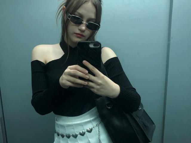 Bubochka with bare shoulders, black off-shoulder top, white pleated skirt, black handbag, and sunglasses, taking a mirror selfie in an elevator, showing her tits.
