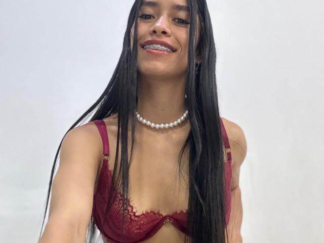 LaurentRay with long black hair, red lace bra, pearl necklace, smiling with metal braces, showing small tits, white background.