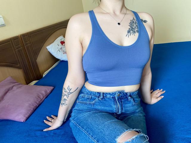 Swoiia with pale skin, blue tank top, showing her tits, tattoo on left arm and right shoulder, blue jeans, sitting on blue bed, pink pillow in background.