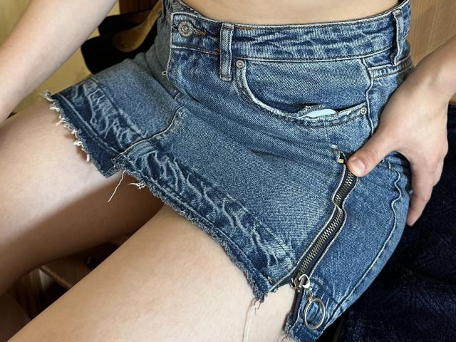 JoannaSia's denim skirt with frayed edges and a zipper on the side, sitting with her hand on her thigh, showing her bare legs and slightly tanned skin.