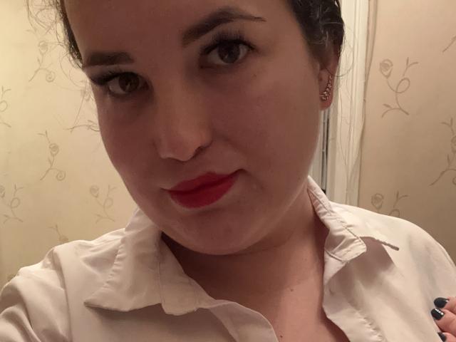 VladaCandy has red lips, light skin, and dark hair in a bun, wearing a white button-up shirt, with small earrings and a slight smile. Her tits are not visible.
