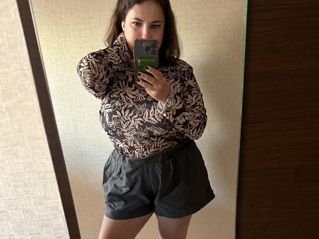 VladaCandy with brown hair, wearing a patterned blouse and black shorts, taking a mirror selfie with a green phone, showing her tits and ass in the reflection.
