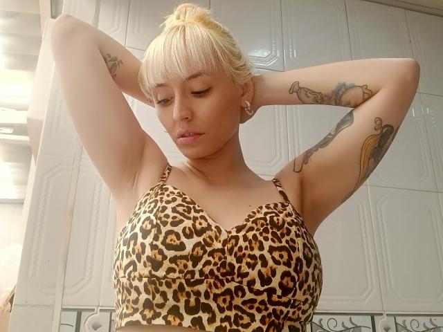 SaraleePoison with blonde hair, leopard print top, arms raised, showing tattoos on both arms. She has small nipples and a slim, attractive build. Her eyes are closed.