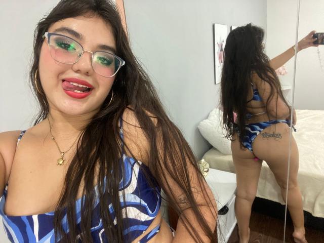 VictoriaSaens has long dark hair, green glasses, and a blue patterned bikini showing her tits and ass with a tattoo on her butt. She's sticking out her tongue in a mirror selfie.