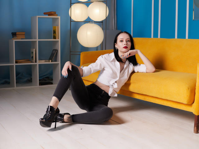 EliseVance with black hair, red lipstick, white shirt tied at the front, black pants, and black high heels, sitting on the floor in front of a yellow couch.