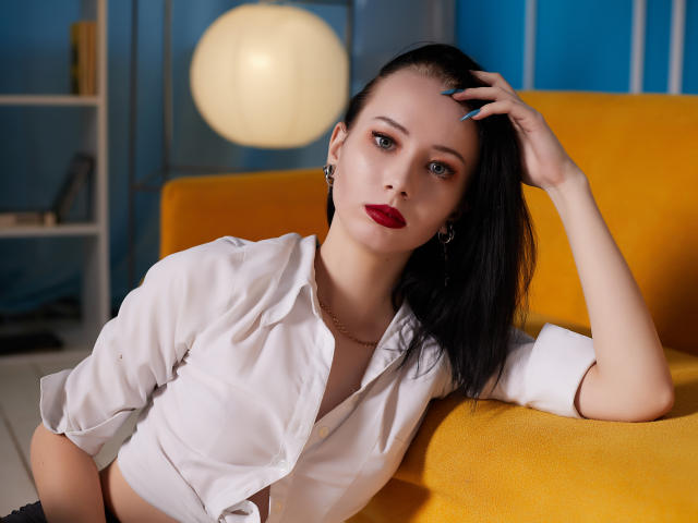EliseVance with black hair, red lipstick, and white button-up shirt, leaning on yellow sofa, hand in hair, blue nails, gold earrings, and subtle necklace, looking at camera.