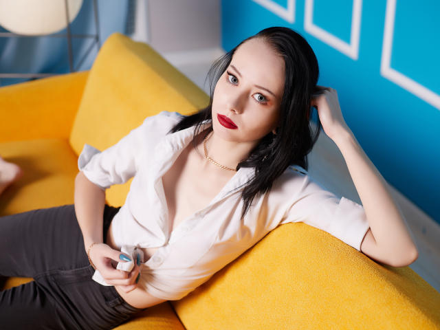 EliseVance with black hair, red lipstick, white blouse, and black pants, lounging on a yellow couch, holding a small device, looking at the camera with a seductive gaze.