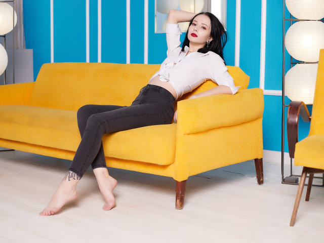 EliseVance is lounging on a yellow couch, wearing a white tied-up shirt and black jeans, with her ass slightly raised, bare feet, and black hair.