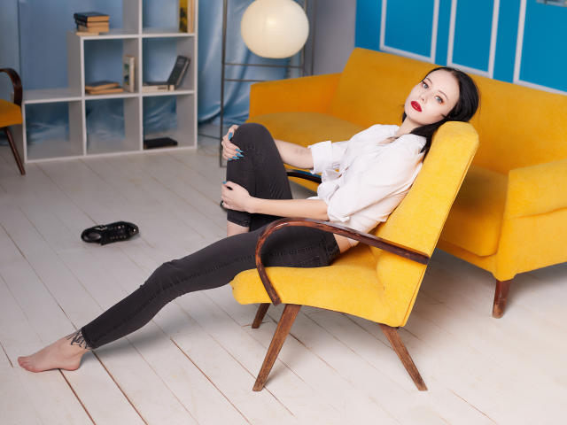 EliseVance with black hair, red lipstick, white blouse, black jeans, and yellow armchair, legs spread, holding jeans, barefoot, black flats on floor, tattoo on right ankle.