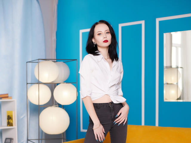 EliseVance has pale skin, black hair, red lipstick, tied white shirt, black pants, standing, hand on hip, looking at camera, room with blue walls, lamp with white spheres.