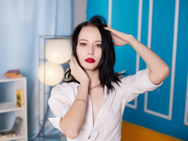 EliseVance with black hair, red lipstick, white shirt, hands in hair, looking at camera, blue wall, white lamp, wooden floor, subtle jewelry, soft light, slightly annoyed expression.