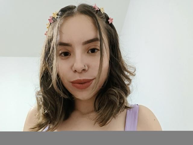 ValentinaSwallow with wavy brown hair, flower crown, purple tank top, nose ring, and pouty red lips, looking down at the camera. Her tits are visible.