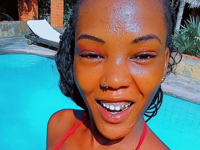 IrahHotie tem uma pele marrom escura e brilhante, um piercing no nariz e um sorriso feliz que mostra dentes brancos. Ela está vestindo um top de biquíni vermelho e está parada ao lado de uma piscina azul vibrante..