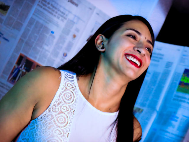 ZaraBeicker, smiling with red lipstick, black hair, white lace sleeveless top, visible ear tattoo, blue light, looking up. Boobs, small to medium, partially visible. tits.