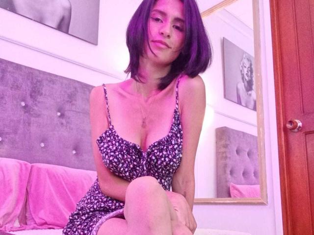 JasminGarcia with purple hair, wearing a floral dress, sitting on a bed, showing small tits, pink bedroom, looking at camera, smiling, arms crossed, knee up.