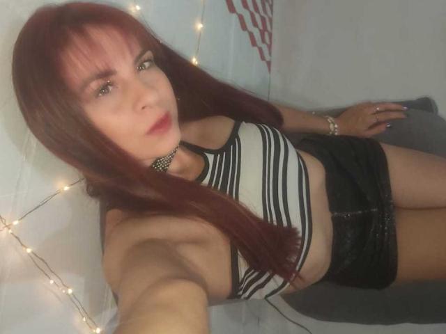 CristalSwt has red hair, wearing a striped tank top, black leather shorts, and a choker, with fairy lights in the background, holding a selfie. Her tits are visible.