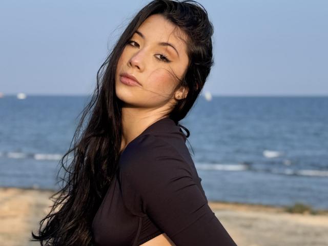 ScarlletRays with long black hair, wearing a black top, looking over her shoulder, showing her bare ass, by the blue ocean. Her face is slightly flushed, hair in wind.